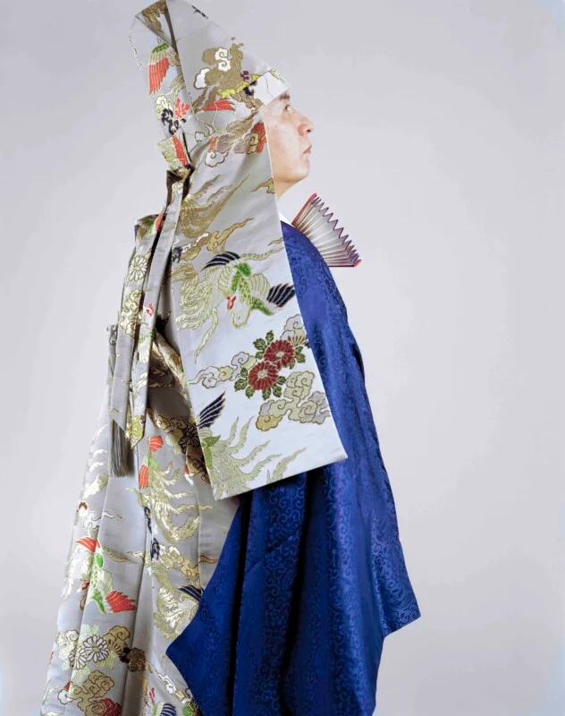 A person in profile wears a traditional Japanese ceremonial outfit, including a tall, patterned silver headpiece and a deep blue robe, while holding a folded fan near their neck.