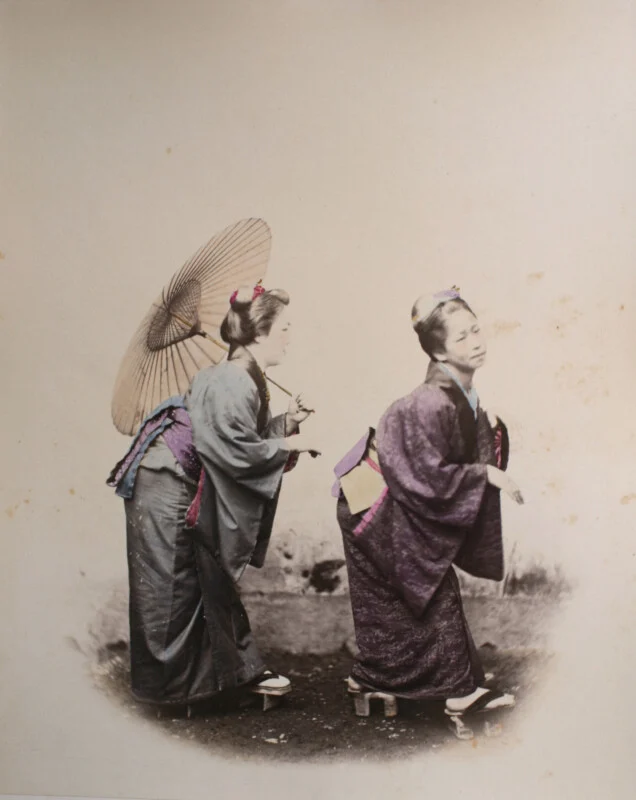 Two women in traditional Japanese kimono and wooden geta sandals stand on a dirt path, one holding a large paper umbrella.