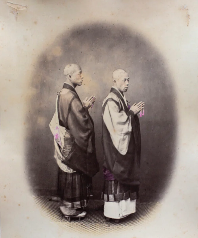 Two Japanese monks with shaved heads stand in profile, wearing traditional robes and holding prayer beads in their hands.