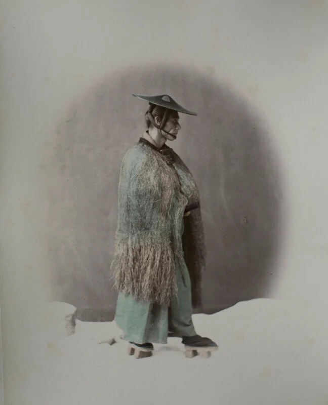 A man in traditional Japanese attire, including a straw rain cape, wide-brimmed hat, and wooden sandals, stands in profile against a plain background.
