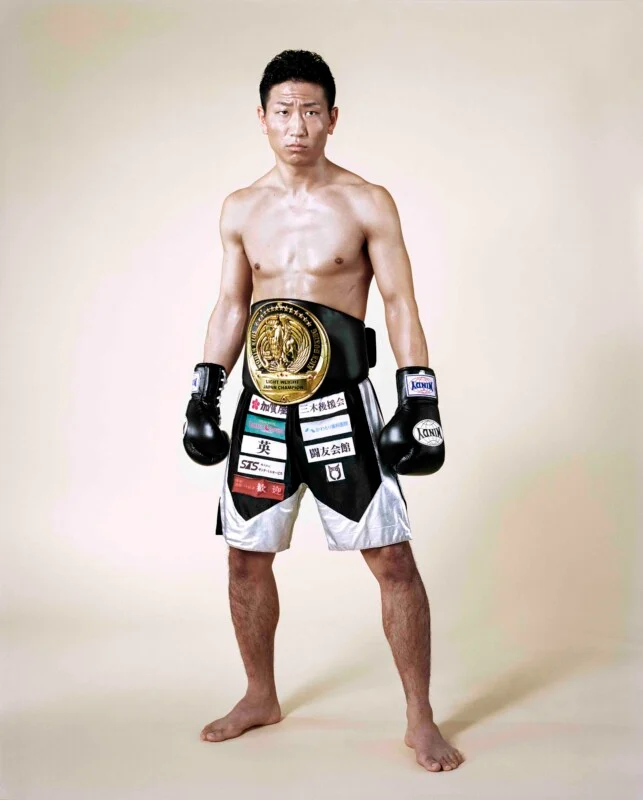 A shirtless male kickboxer stands barefoot against a plain background, wearing black and silver trunks with sponsor patches and a championship belt.