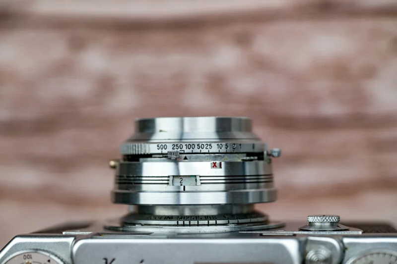 A close-up view of a vintage camera lens barrel featuring shutter speed markings from 500 to 1, a focus ring with distance measurements in feet, and a red X symbol.
