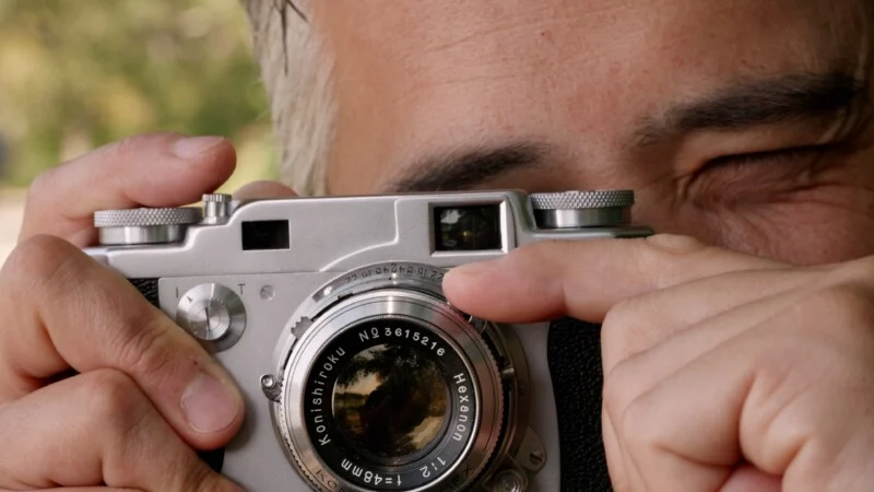 A close-up view of a person holding a vintage silver film camera to their eye, with their finger resting on the lens aperture ring.