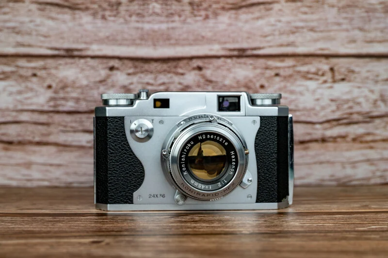 A vintage Konishiroku Hexanon 35mm film camera sits centered on a wooden surface against a light wood-grain background.