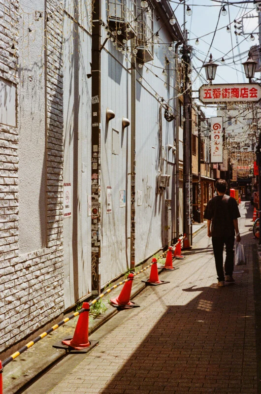 A person walks down a narrow, sunlit alleyway lined with orange traffic cones and a brick wall covered in stickers.
