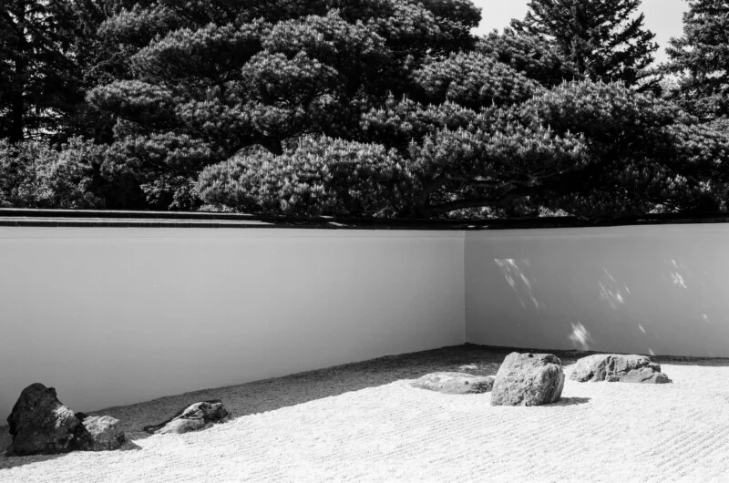 A black and white view of a Japanese rock garden with raked gravel, several large stones, a white wall, and overhanging trees.