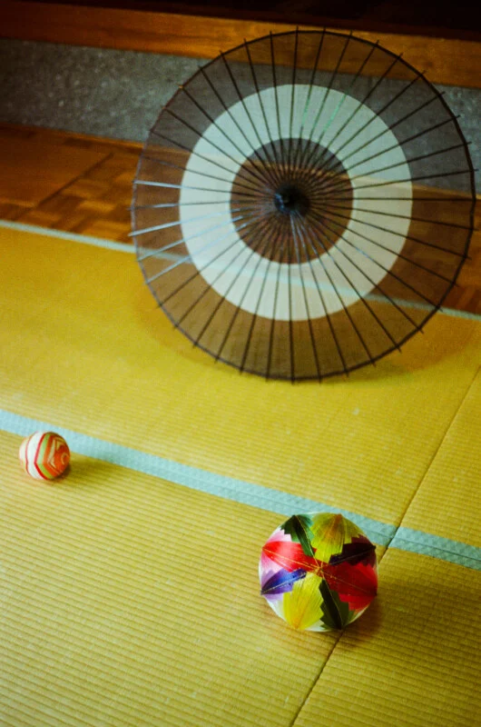 A traditional Japanese paper umbrella stands upright on a tatami mat floor near two colorful, patterned temari balls.