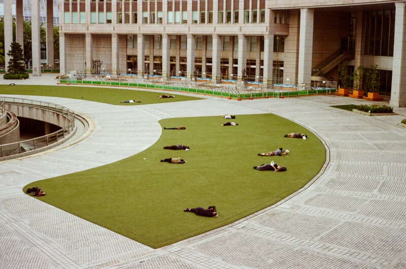 A high-angle view of a curved patch of green lawn in a paved plaza where several people are lying down and resting.