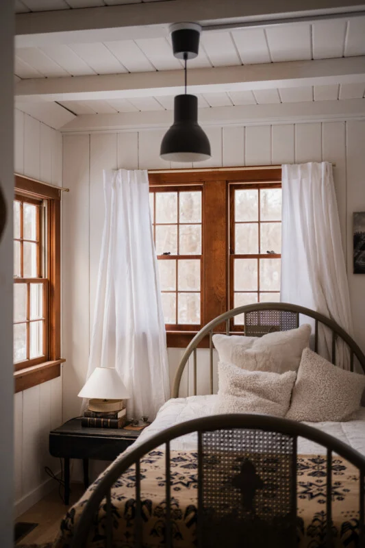 A bedroom features a bed with a patterned blanket and textured pillows, a small black side table with a lamp and books, and windows with white curtains.