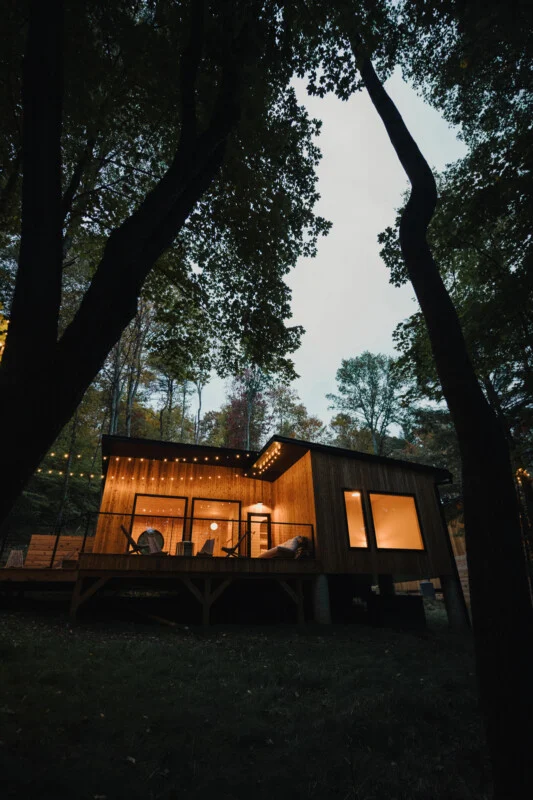 A modern wooden cabin with large windows and a deck sits in a forest at dusk, illuminated by warm string lights.