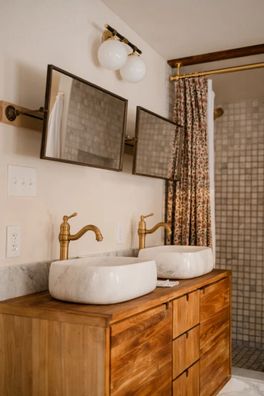 A double vanity with a wooden cabinet, two marble vessel sinks, brass faucets, and two rectangular mirrors mounted on a wooden bar.