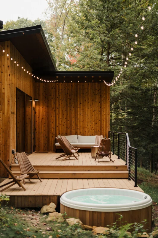 A wooden deck with a hot tub, outdoor seating, and string lights attached to a modern cabin in a wooded area.