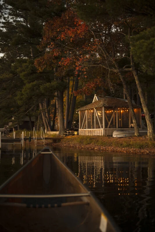A canoe floats on a calm lake toward a screened-in gazebo illuminated by string lights on a wooded shoreline during autumn.