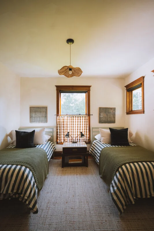 A bedroom features two twin beds with striped bedding and green throws, separated by a small nightstand with a double lamp under a window with a gingham curtain.