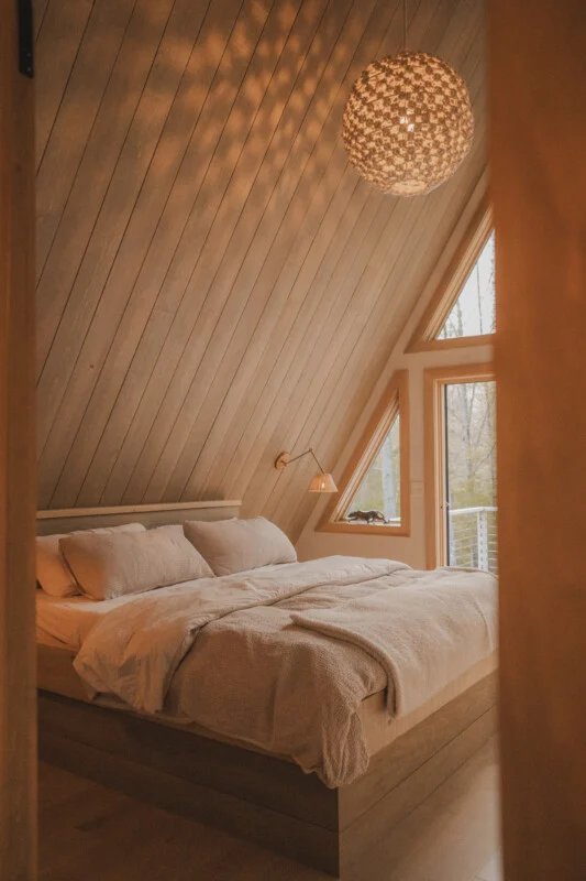 A cozy bedroom in an A-frame cabin features a platform bed with neutral bedding, a textured pendant light, and a window overlooking a forest.