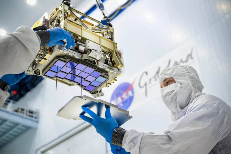 A technician in a cleanroom suit and blue gloves assembles a space instrument with a grid of purple sensors.