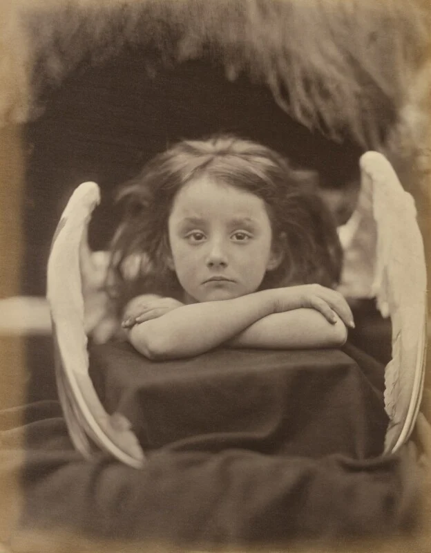 A sepia-toned portrait of a young child with long, dark hair and feathered wings, resting their chin on crossed arms.