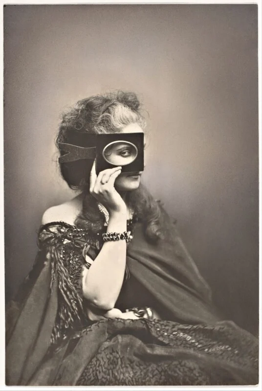 A woman with wavy hair wears an ornate dark dress and holds a rectangular frame with an oval cutout over one eye, revealing her gaze.