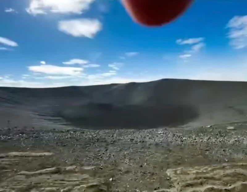 A wide, bowl-shaped volcanic crater with dark, rocky slopes under a bright blue sky with scattered clouds.