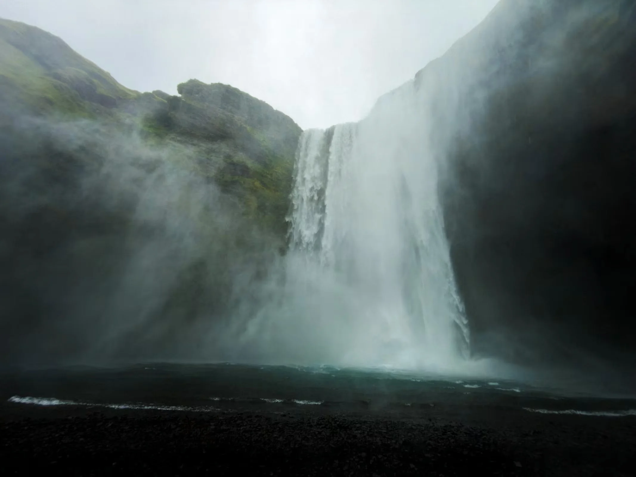 A massive waterfall cascades down a dark, mossy cliff face into a pool of water, creating a thick mist at the base.
