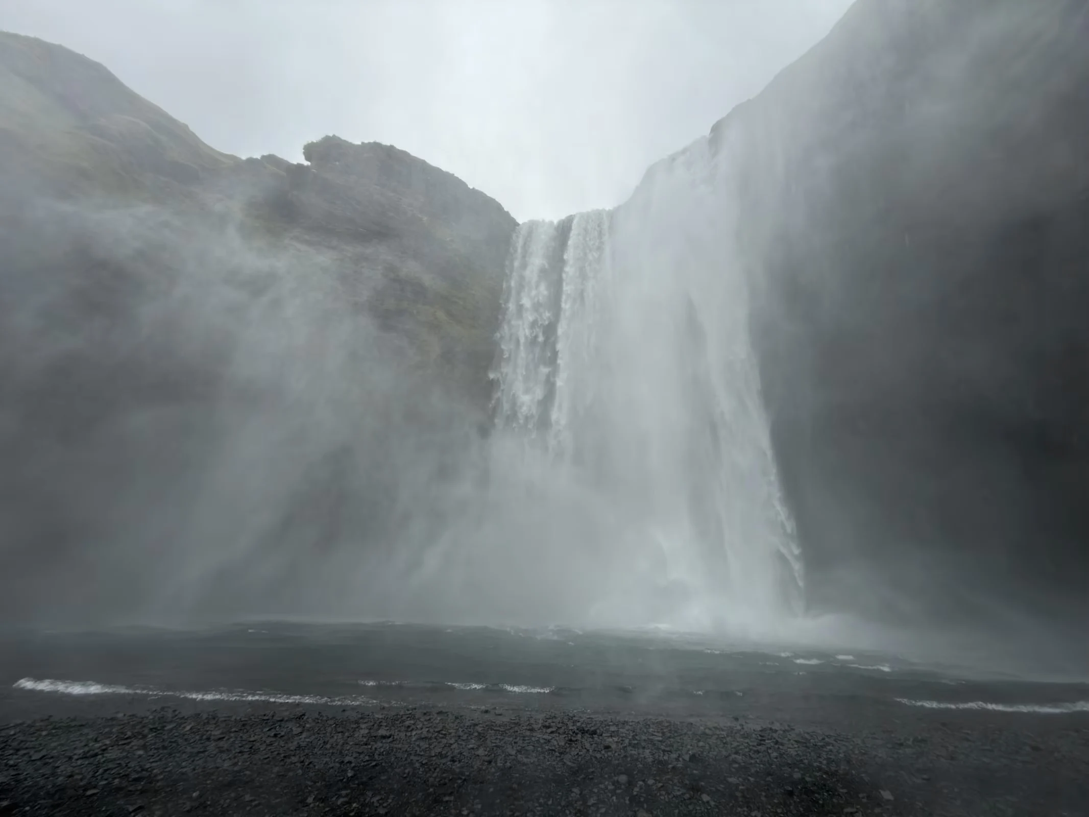 A massive waterfall cascades down a dark, rocky cliff face into a pool, creating a thick mist at the base.