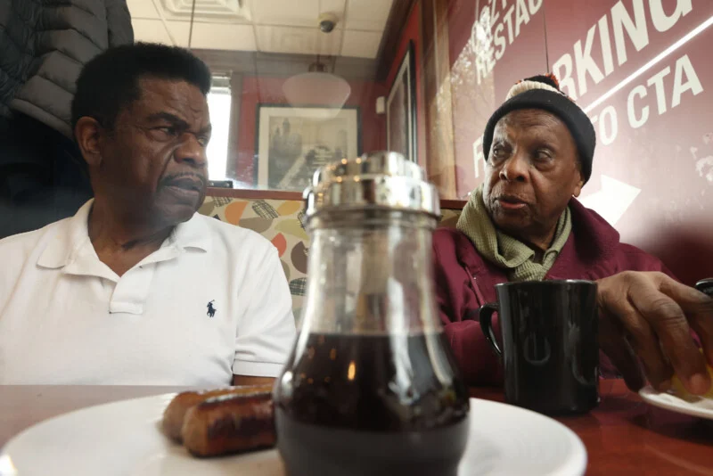 Two older Black men sit at a restaurant table, looking at each other while a syrup dispenser sits in the foreground.