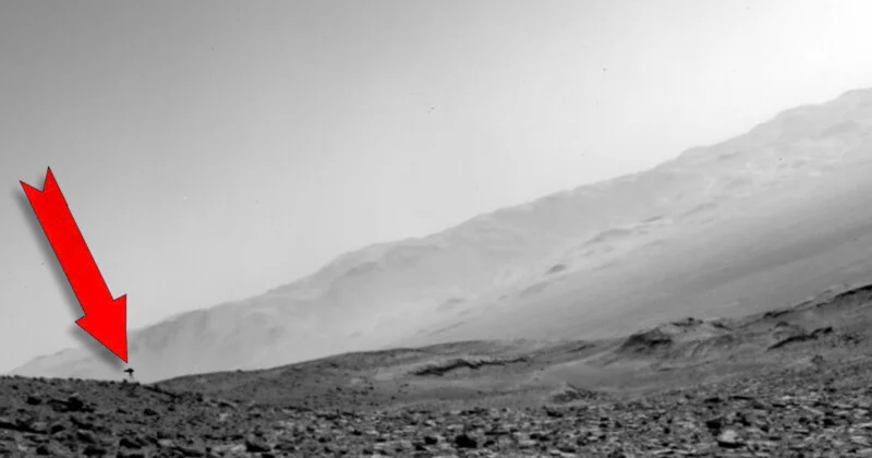 A red arrow points to a small, dark, T-shaped object resting on a rocky, barren landscape beneath a large, sloping hill.