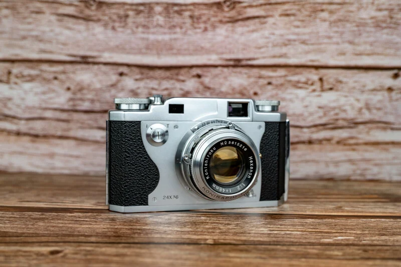 A vintage Konishiroku Hexanon 35mm rangefinder camera sits on a wooden surface against a wood-paneled background.