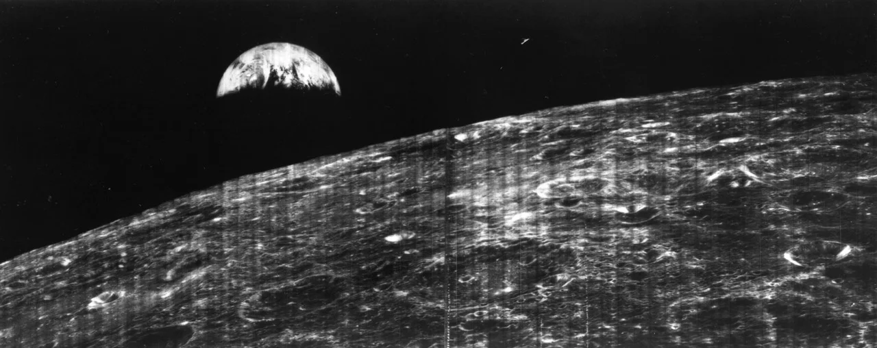 A black-and-white view of the Moon's cratered surface in the foreground with the Earth rising in the dark sky above.