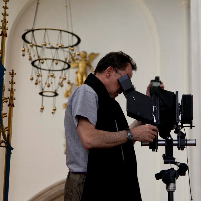 A man in a grey t-shirt and black scarf looks through the viewfinder of a large-format camera mounted on a tripod inside a building.