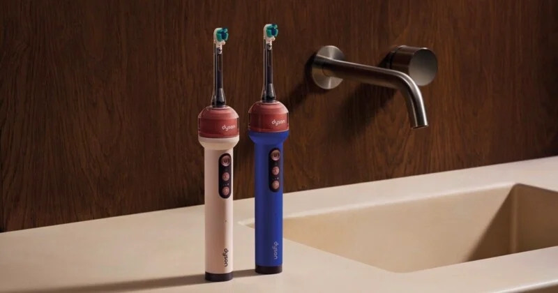 Two Dyson electric toothbrushes, one cream and one blue, stand on a bathroom counter next to a sink faucet.