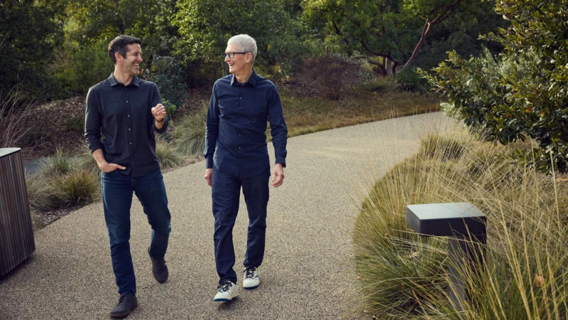 Two men walk along a gravel path in a park, smiling and looking at each other while wearing button-down shirts and jeans.