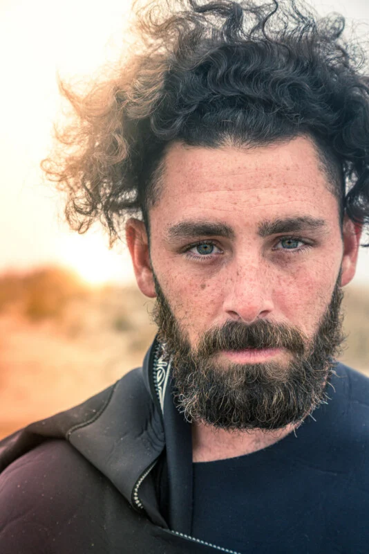A close-up portrait of a man with curly dark hair, a full beard, and freckles, wearing a black wetsuit against a blurred, sunlit background.