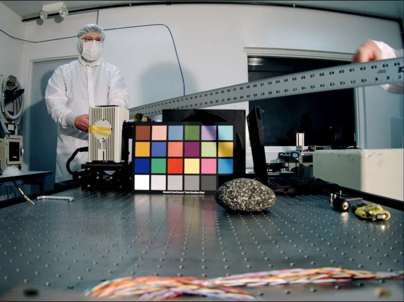 A person in a white cleanroom suit stands behind a table holding a metal ruler, with a color calibration chart and a speckled rock placed on the surface.