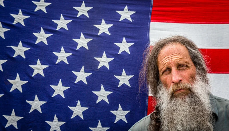 A man with long gray hair and a full beard stands in front of a large American flag.
