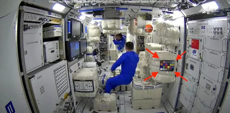 Two astronauts in blue jumpsuits float inside the interior of a space station module, with a color calibration chart attached to a cargo container.