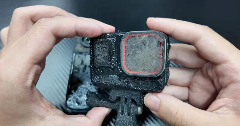 A pair of hands holds a heavily mud-caked action camera with a red-rimmed lens cover.