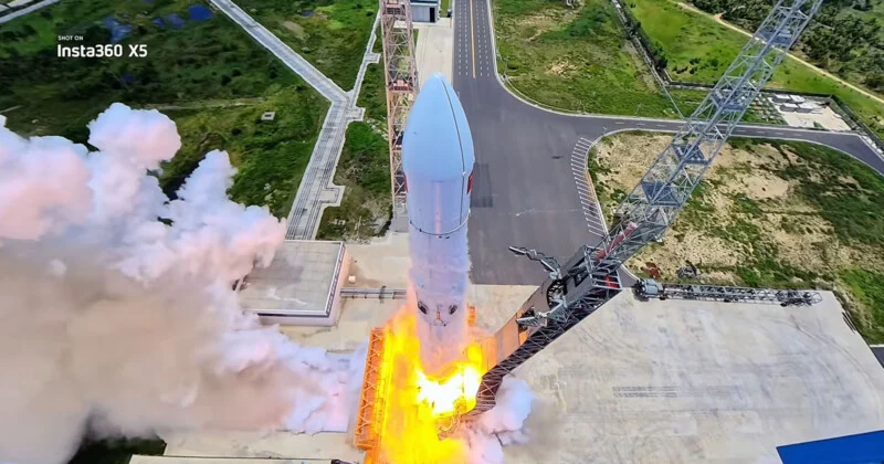 A rocket launches from a pad, surrounded by bright orange flames and large plumes of white smoke, with a tall service tower visible to the right.