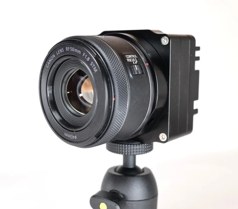 A Canon RF 50mm F1.8 STM lens attached to a black, box-shaped camera body mounted on a small tripod head against a white background.