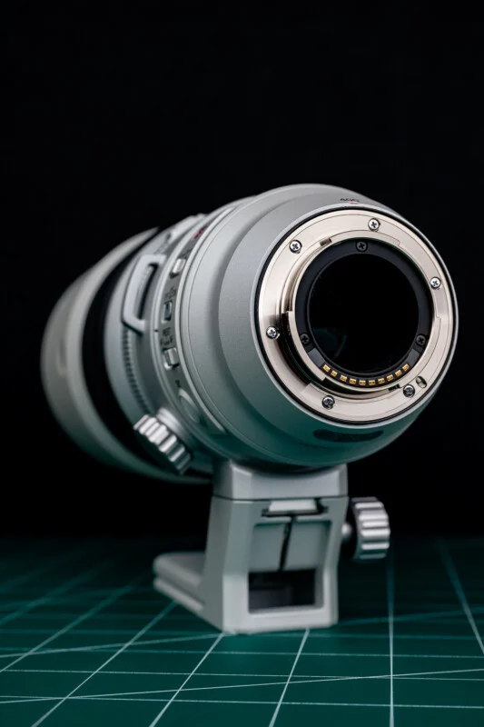 A silver telephoto camera lens sits upright on a green cutting mat against a black background, showing its rear mount and electronic contact pins.
