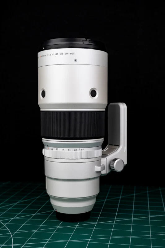 A silver telephoto camera lens stands vertically on a green cutting mat against a black background.
