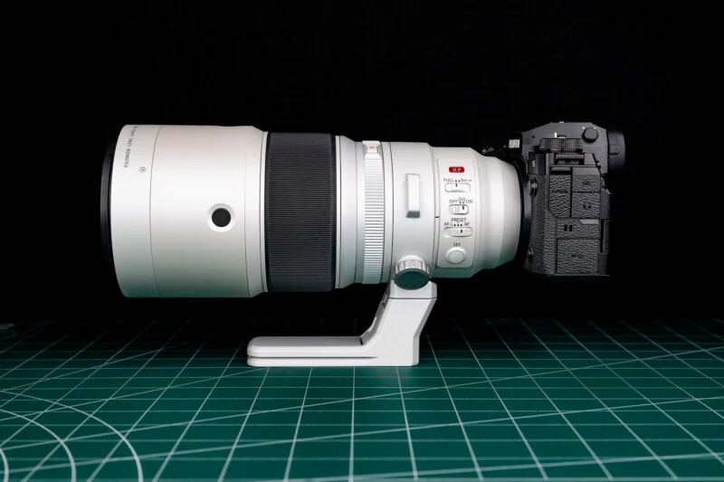 A white telephoto lens attached to a black mirrorless camera body, resting on a green cutting mat against a black background.