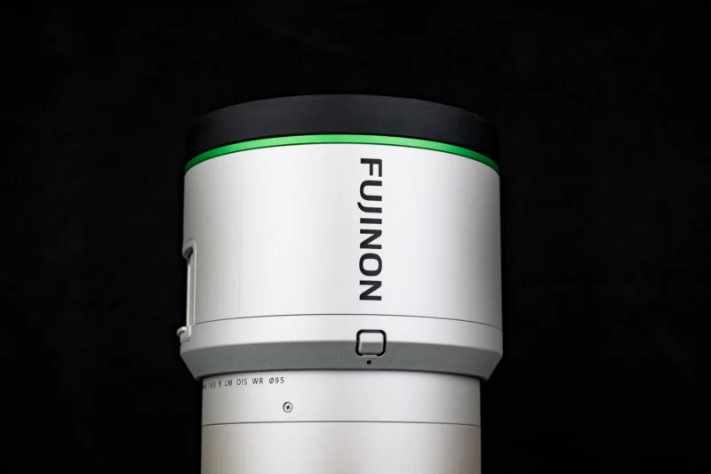 A close-up view of a silver Fujinon camera lens against a black background, featuring a green accent ring and a visible focus button.
