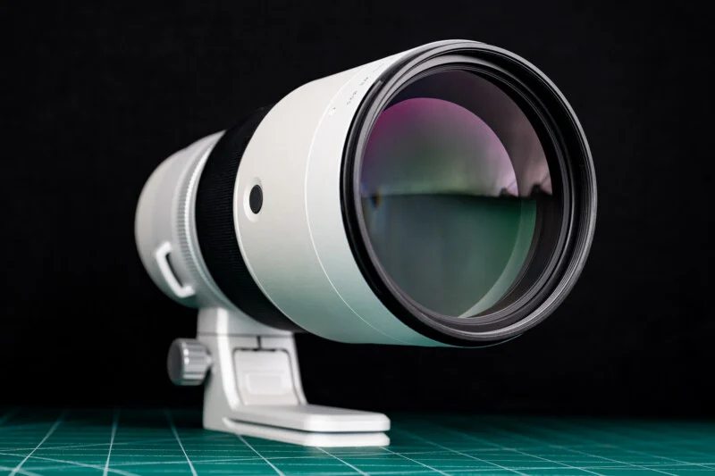 A white telephoto camera lens with a black focus ring sits on a green cutting mat against a black background.