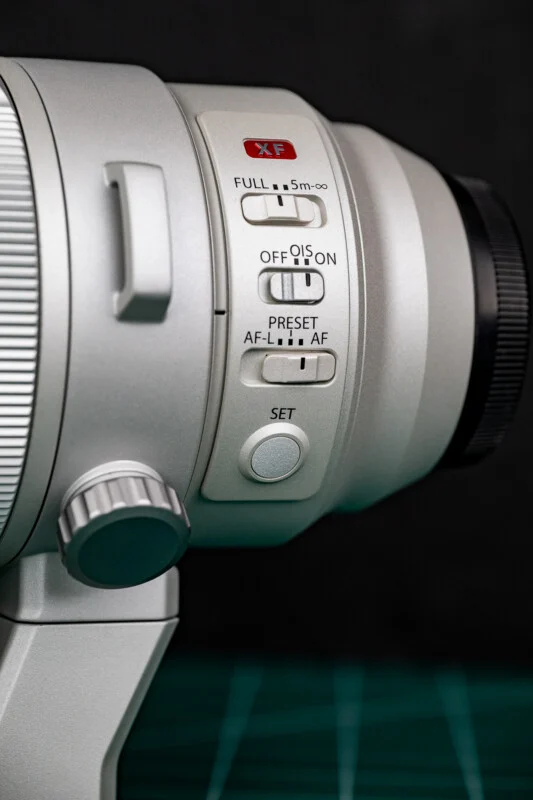 A close-up of a silver camera lens barrel featuring control switches for focus range, optical image stabilization, and autofocus, along with a circular "SET" button.