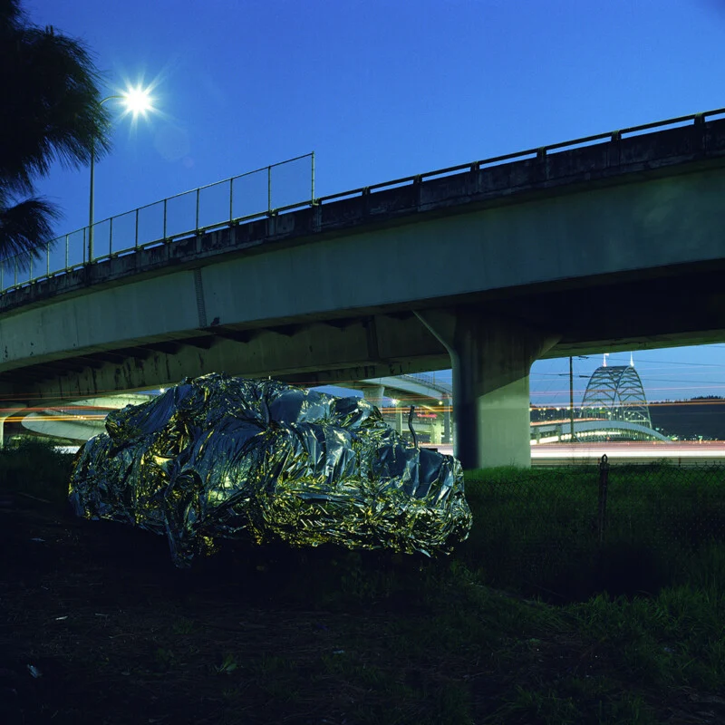 A car covered in a crinkled, reflective silver emergency blanket sits under a highway overpass at dusk, with a bridge visible in the distance.