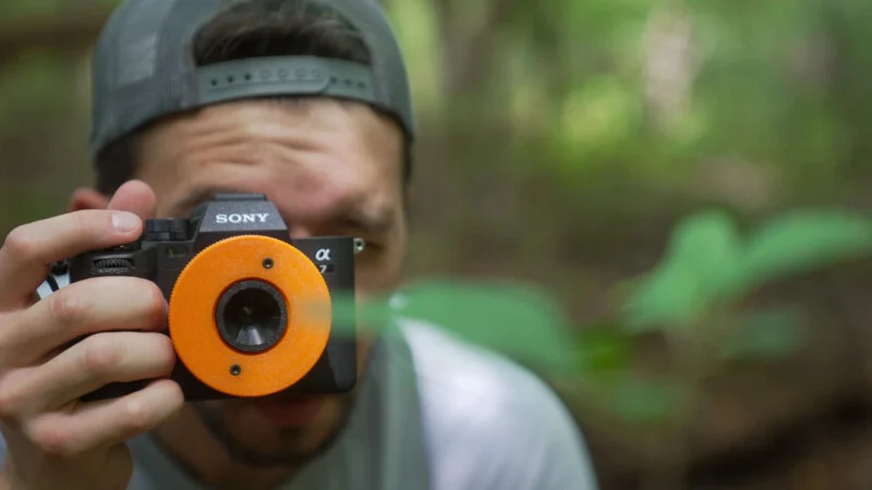A person wearing a baseball cap holds a camera with a bright orange, circular 3D-printed lens cap in front of their face.