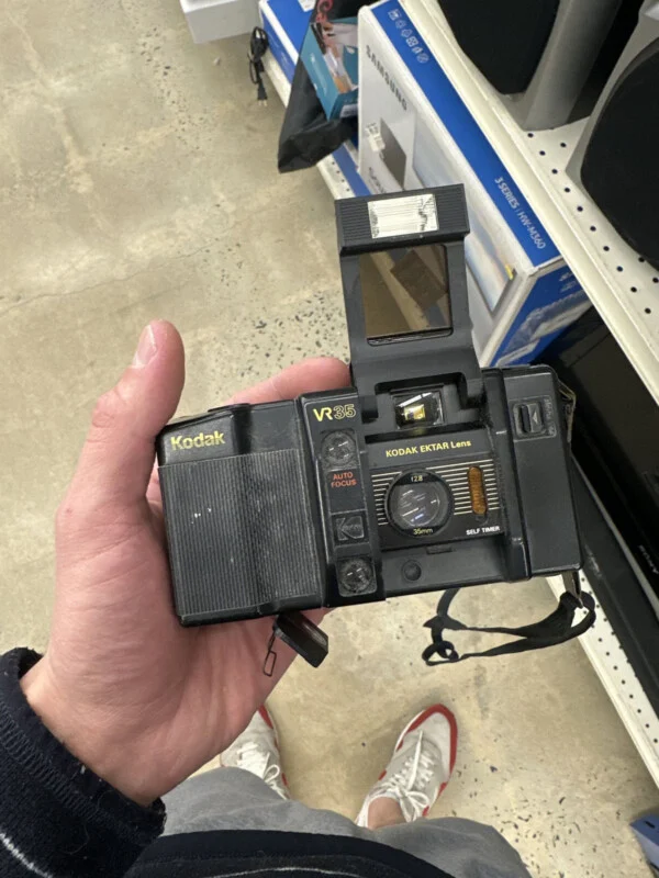 A hand holds a black Kodak VR35 film camera with a raised flash unit in a retail store aisle.