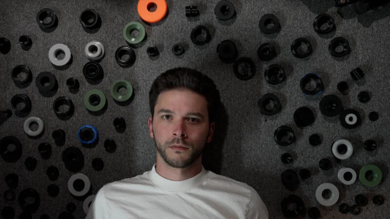 Een man met een baard en een wit T-shirt ligt op een grijze tapijtvloer, omringd door tal van ronde plastic ringen en onderdelen.