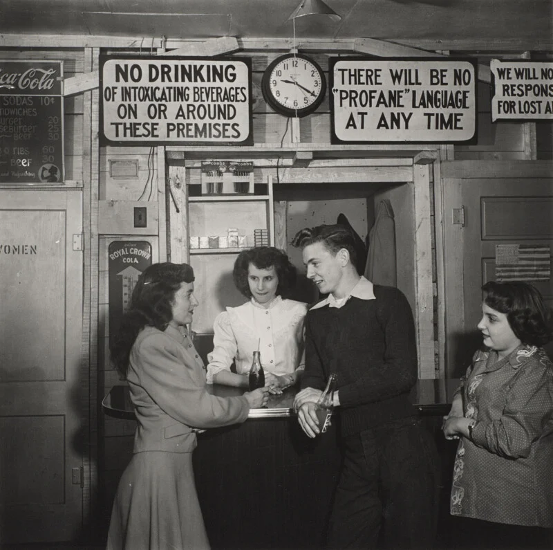 Four young people stand around a counter in a room with signs prohibiting alcohol and profanity, while a menu lists food prices.